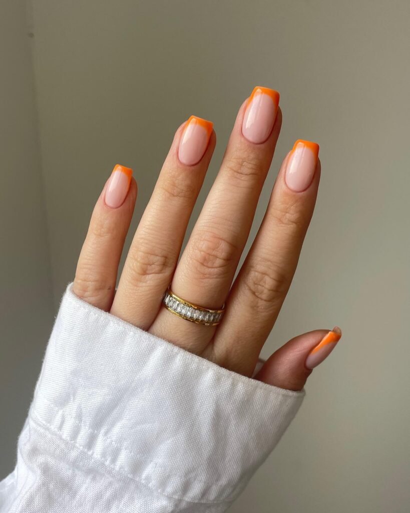 Hand with orange French manicure, wearing a diamond ring, against a neutral background.