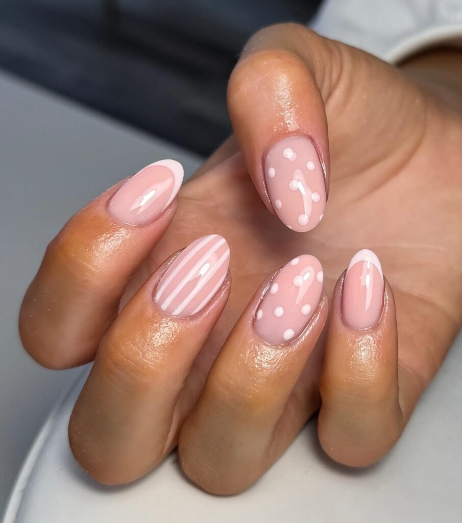 Pink manicure with polka dots, stripes, and French tips on oval-shaped nails, showcasing trendy nail art designs.