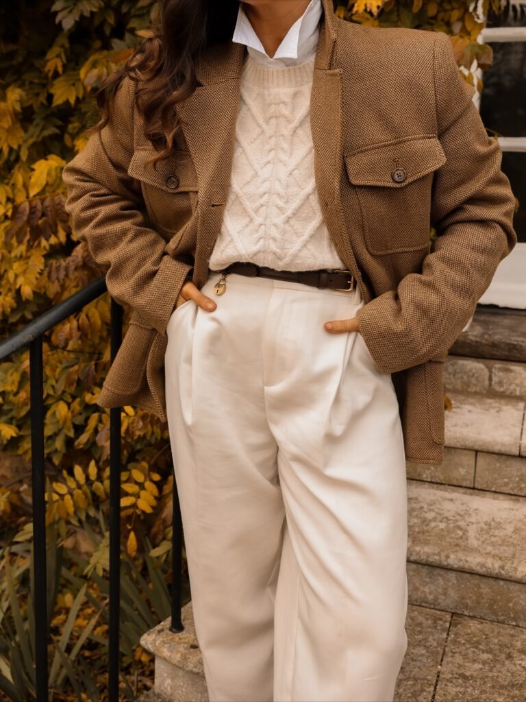 Woman in fall outfit with brown jacket and white pants, standing outdoors by steps with autumn leaves.