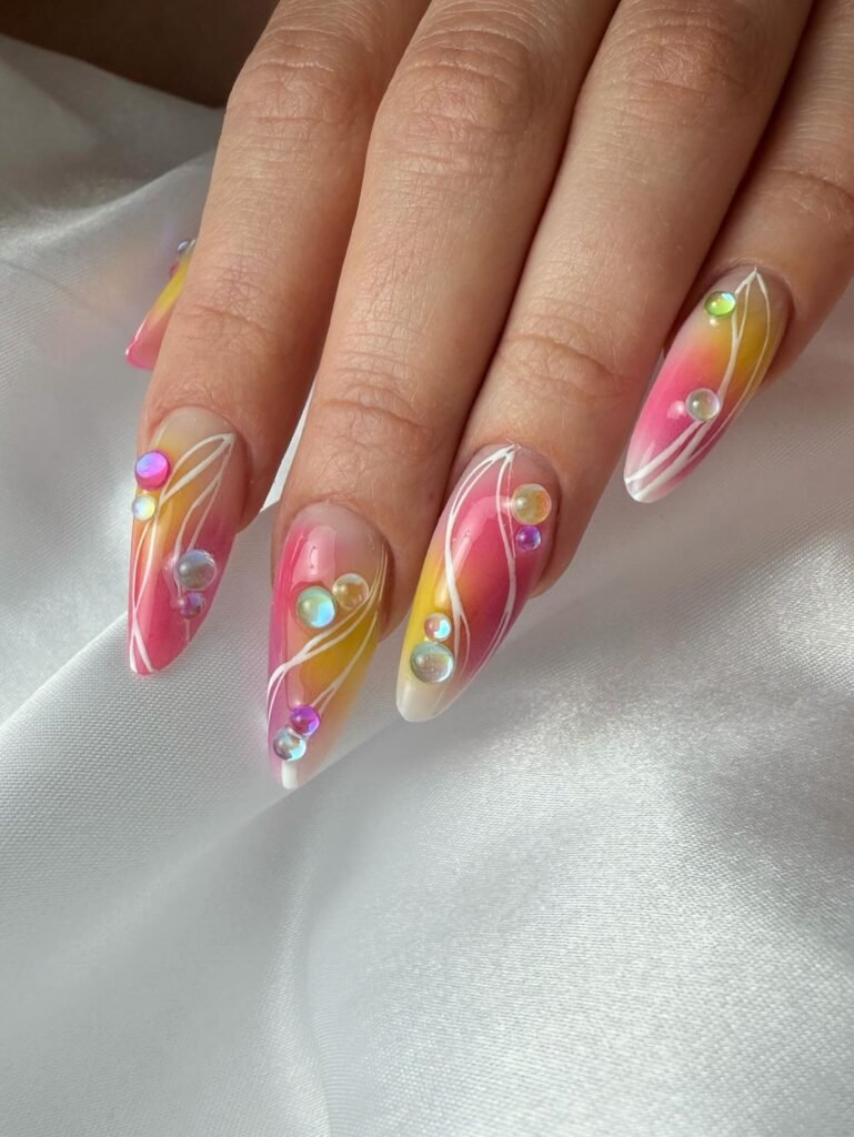 Colorful gradient nail art with rhinestone accents on long, pointed nails, resting on a white silk background.