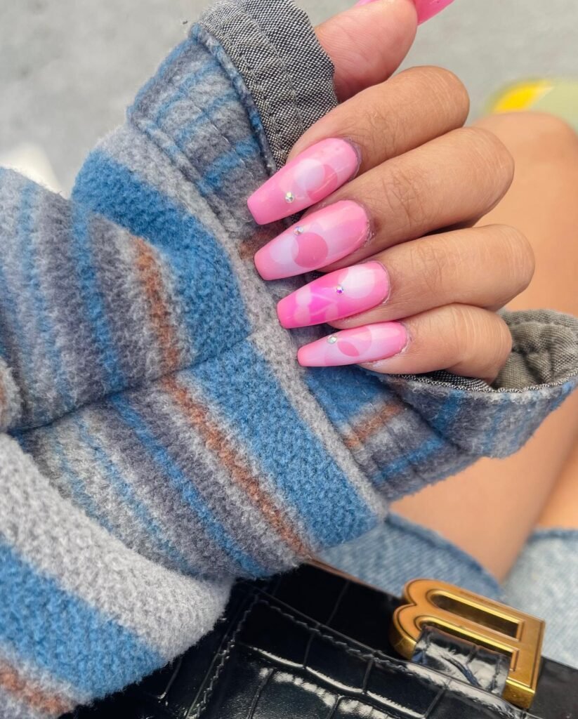 Pink and white abstract nail art with rhinestones on a hand in a blue striped sleeve, holding a black leather purse.