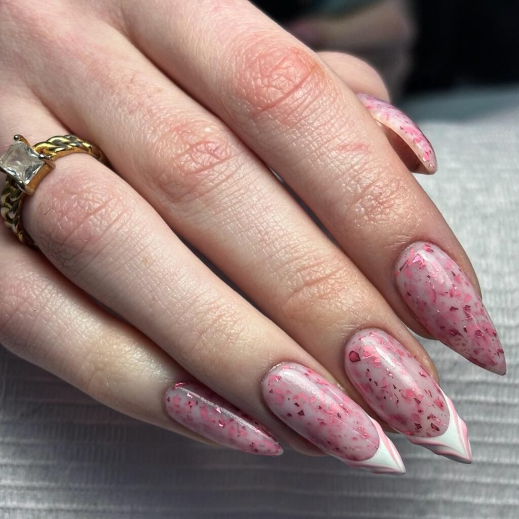 Close-up of hand with pink speckled almond-shaped nails and gold ring, showcasing trendy nail art design.