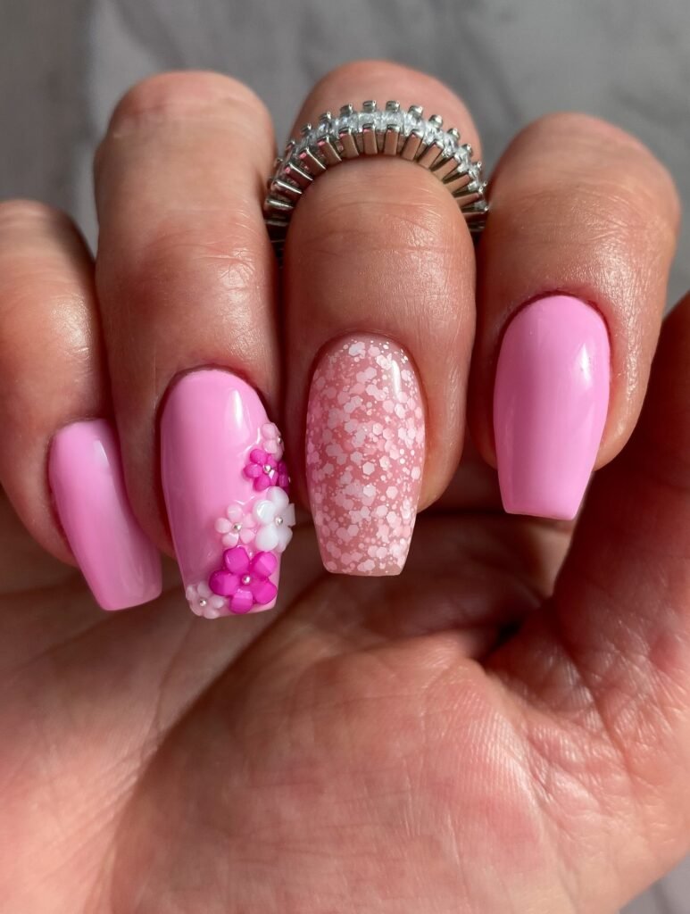 Pink floral nail art with 3D designs and glitter accent, complemented by a stylish silver ring.