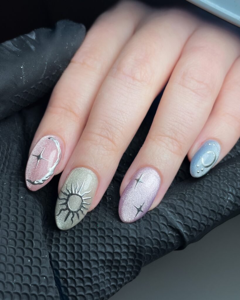 Shimmery celestial nail art with sun, moon, and star designs on pastel colors over black textured fabric.