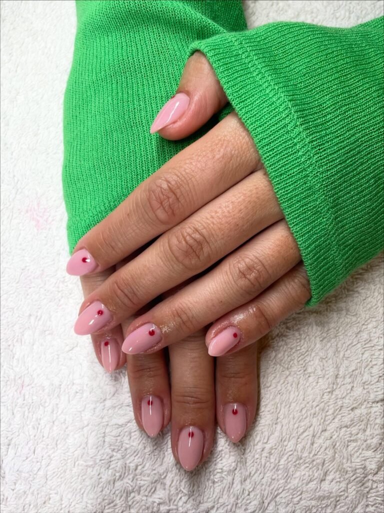 Pink almond nails with red dot designs on a hand wearing a green sweater sleeve, resting on white fabric.