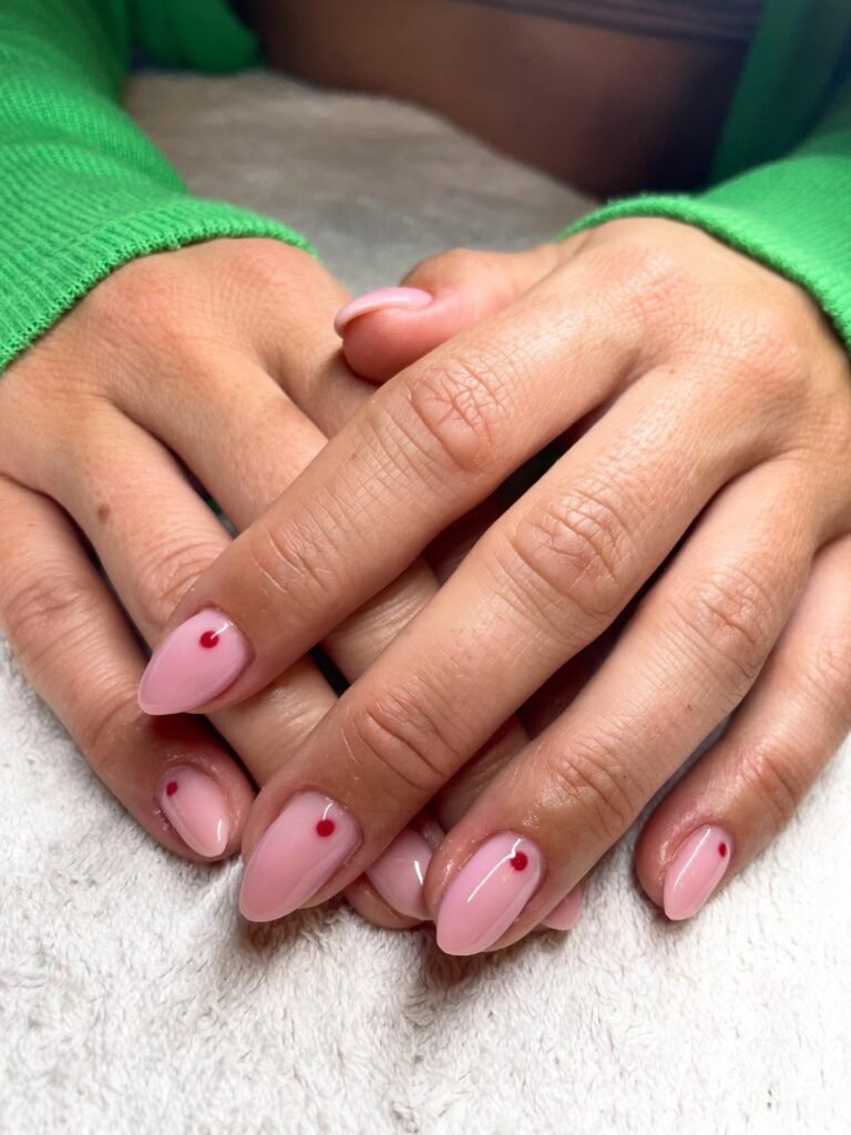 Hands with pink nail polish featuring red dot accents, against a green sweater background.
