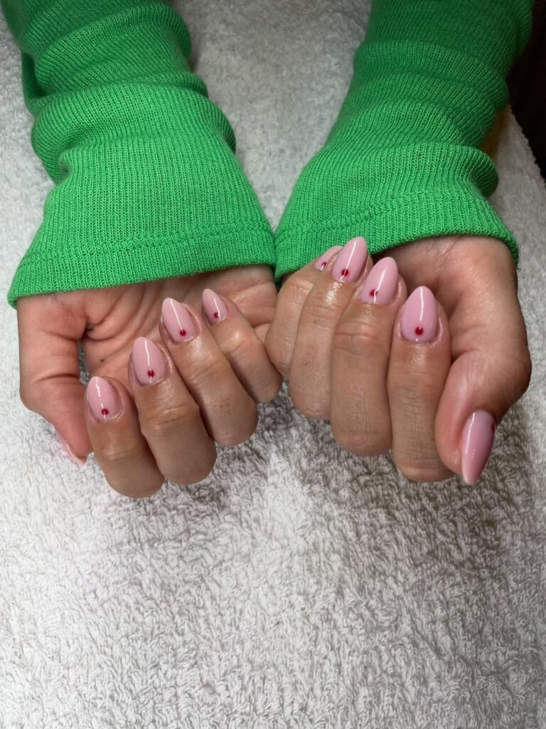 Pink almond nails with red heart design and a green sweater on a white textured background.