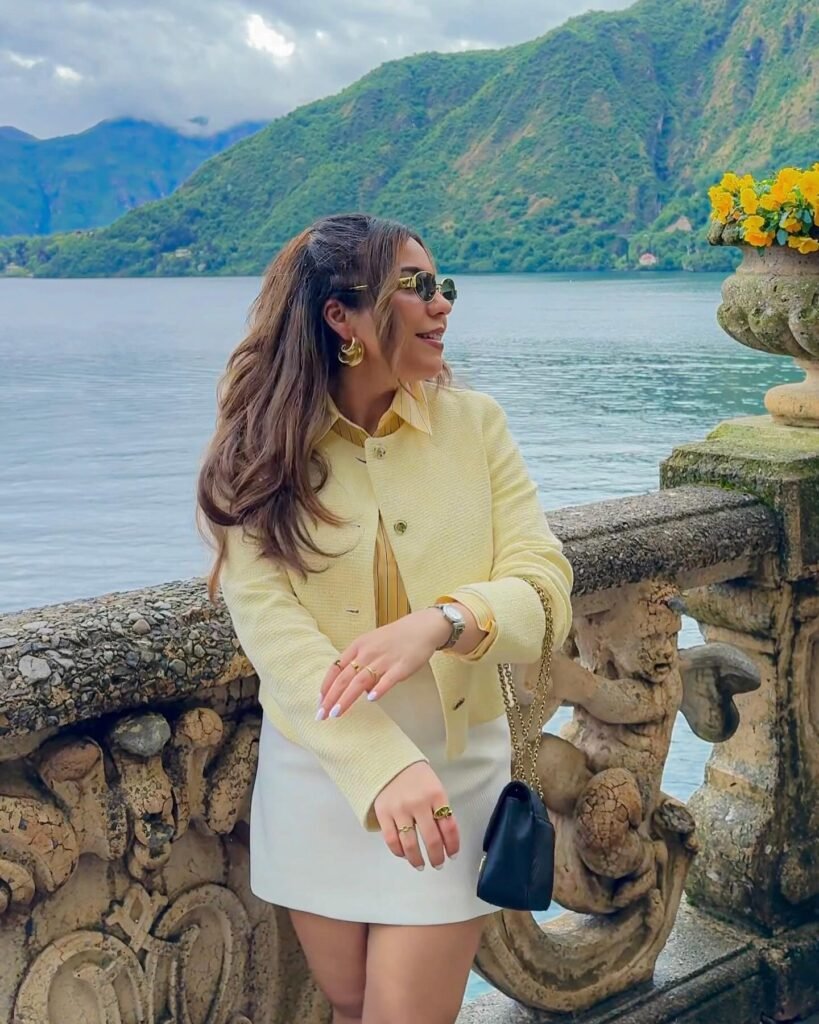 Woman in yellow jacket enjoys scenic lakeside view with mountains, wearing sunglasses and carrying a black bag.