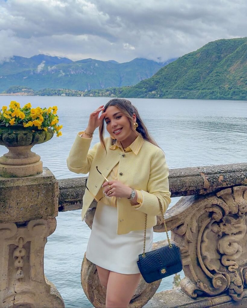 Woman in yellow jacket smiling by scenic lake and mountains, holding sunglasses near stone railing with yellow flowers.