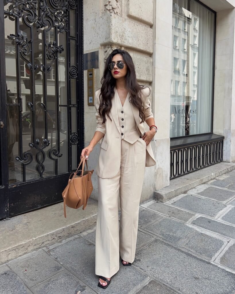Woman in a stylish beige pantsuit with sunglasses and a brown bag, standing on a city sidewalk.