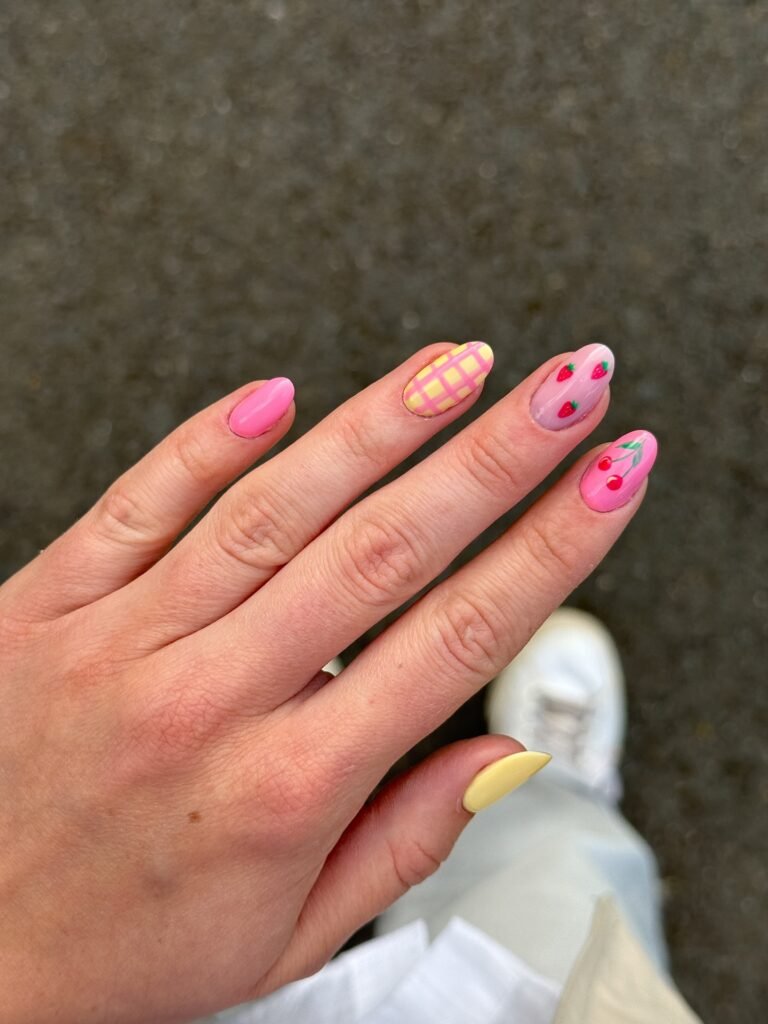 Hand with colorful, playful nail art including pink, yellow, and fruit designs against a blurred background.