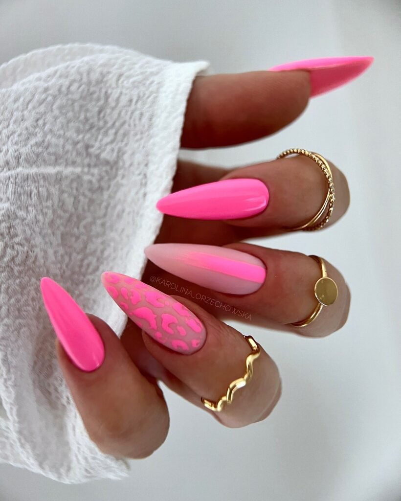Bright pink stiletto nails with leopard print accent, adorned with gold rings against a textured white fabric.