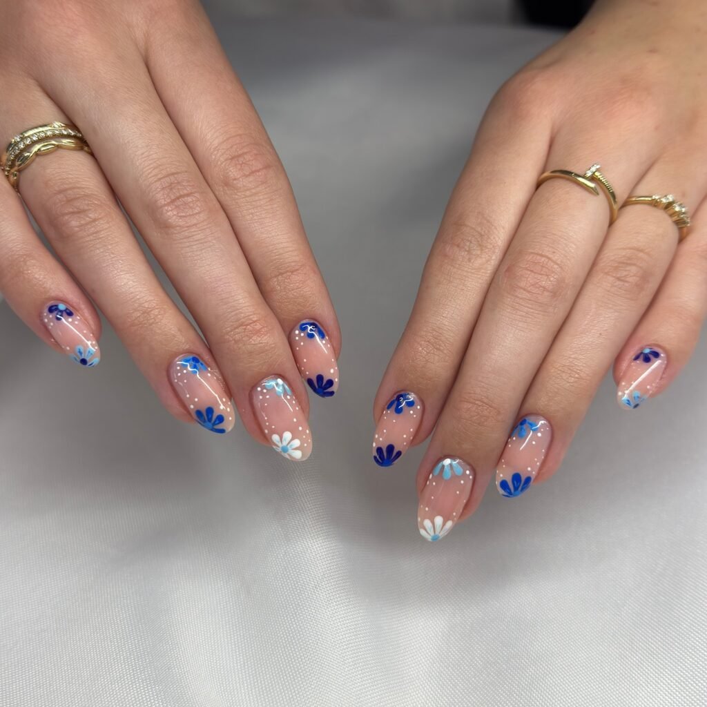 Manicured nails with blue and white floral designs on a nude base, featuring elegant rings.