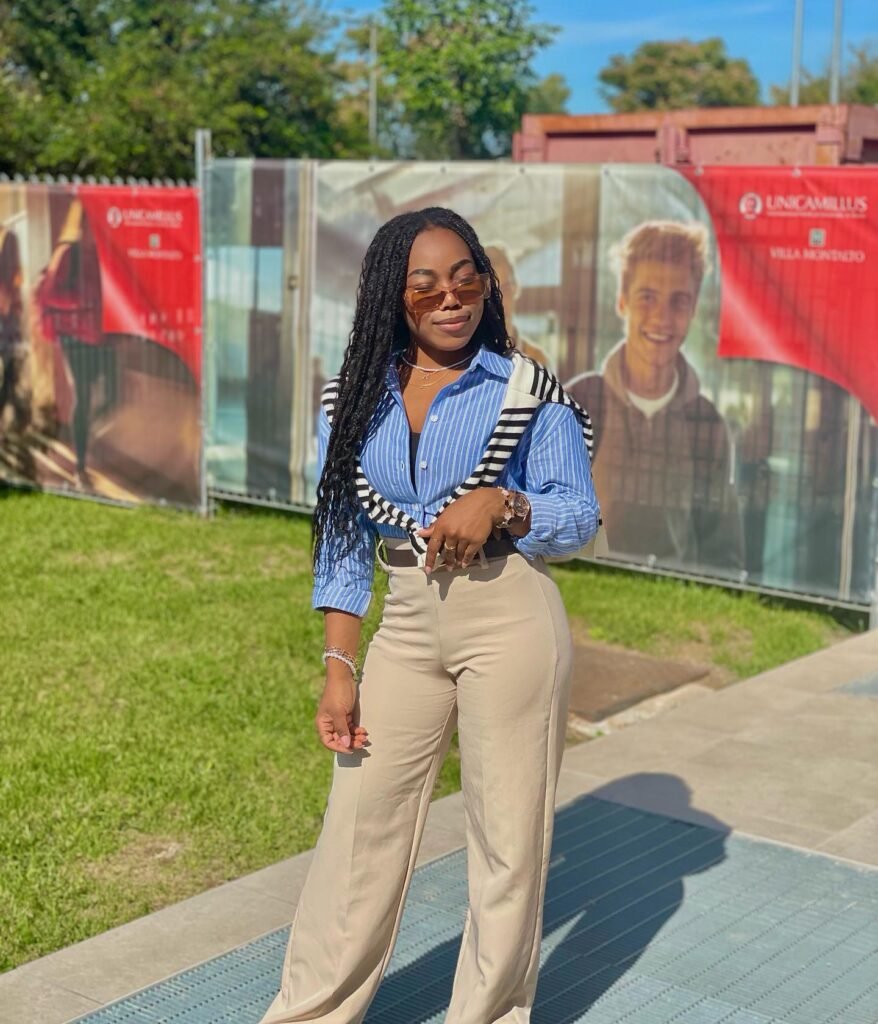 Woman in stylish outfit standing outdoors, wearing sunglasses, blue striped shirt, and beige pants on a sunny day.