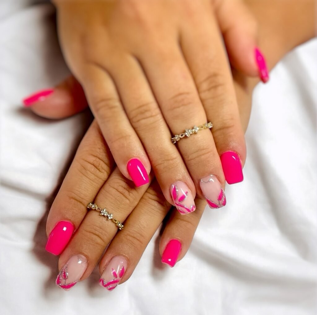 Bright pink nails with white and silver floral accents; gold rings on fingers for an elegant manicure.