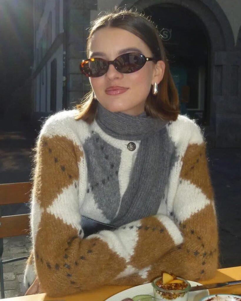 A woman wearing sunglasses and a cozy sweater sits at an outdoor cafe, enjoying a sunny day.