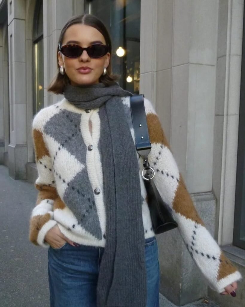 Fashionable woman in argyle cardigan and scarf, wearing sunglasses and jeans, walks on city street.