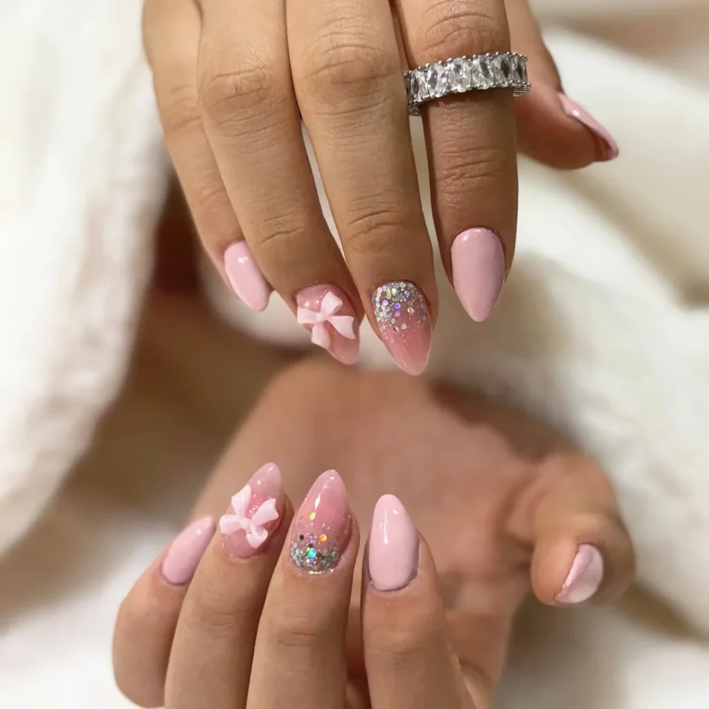 Elegant pink nails with glitter and a 3D bow design, featuring a sparkling ring. Perfect manicure inspiration.
