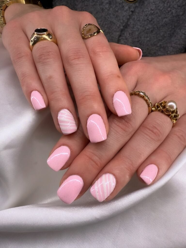 Pink manicure with stripes on a hand wearing gold rings, displayed on white satin fabric.