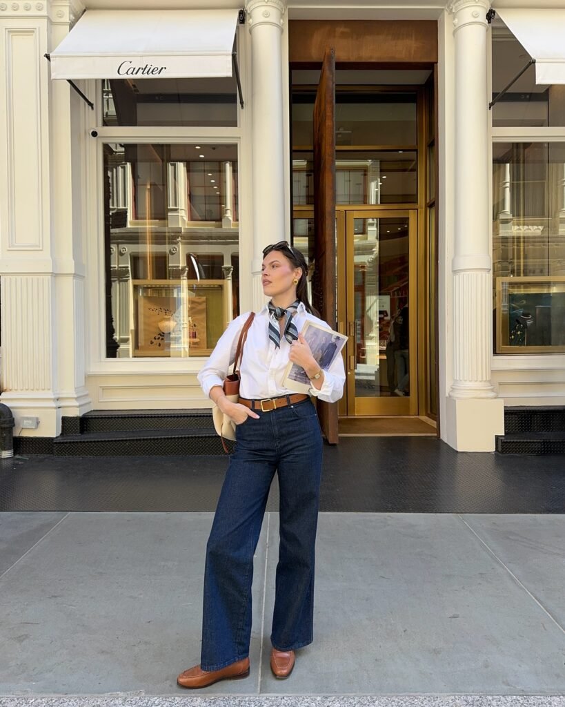 Stylish woman in jeans and white shirt stands confidently outside a luxury store, holding a magazine.