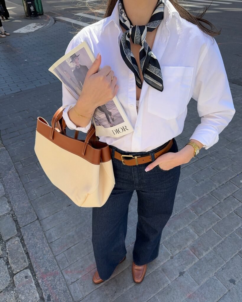 Fashionable woman with scarf, white shirt, holding a magazine, beige tote, and wearing dark jeans and brown loafers.