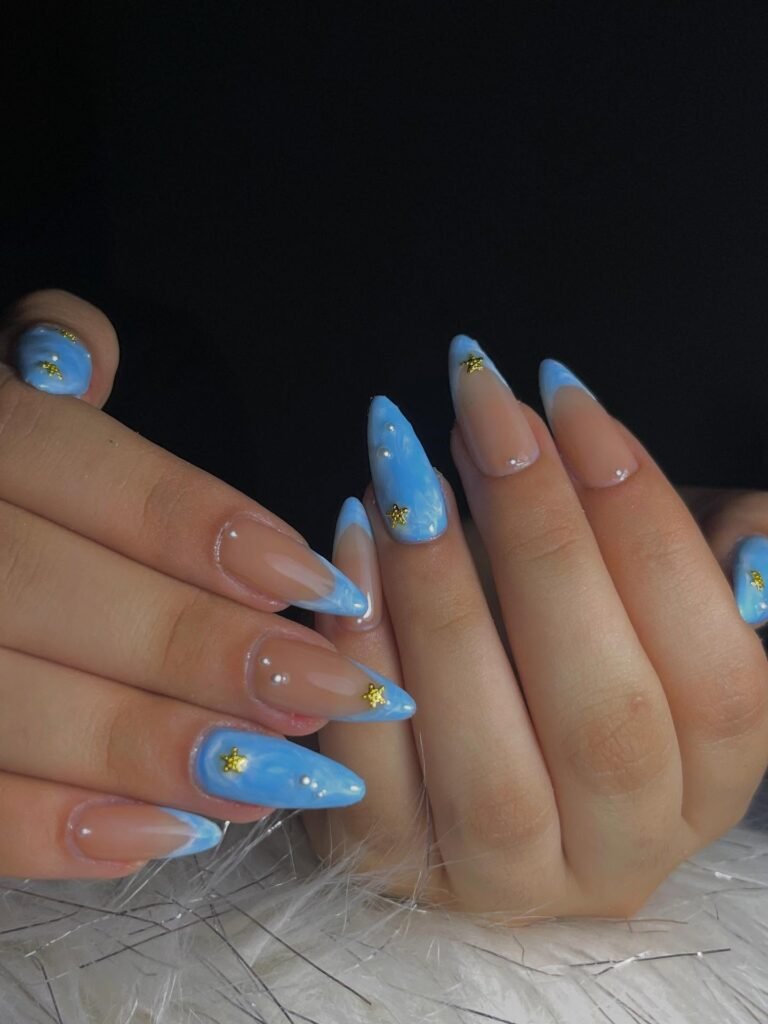 Elegant stiletto nails with blue and nude design, adorned with tiny golden stars and pearls on white fur background.