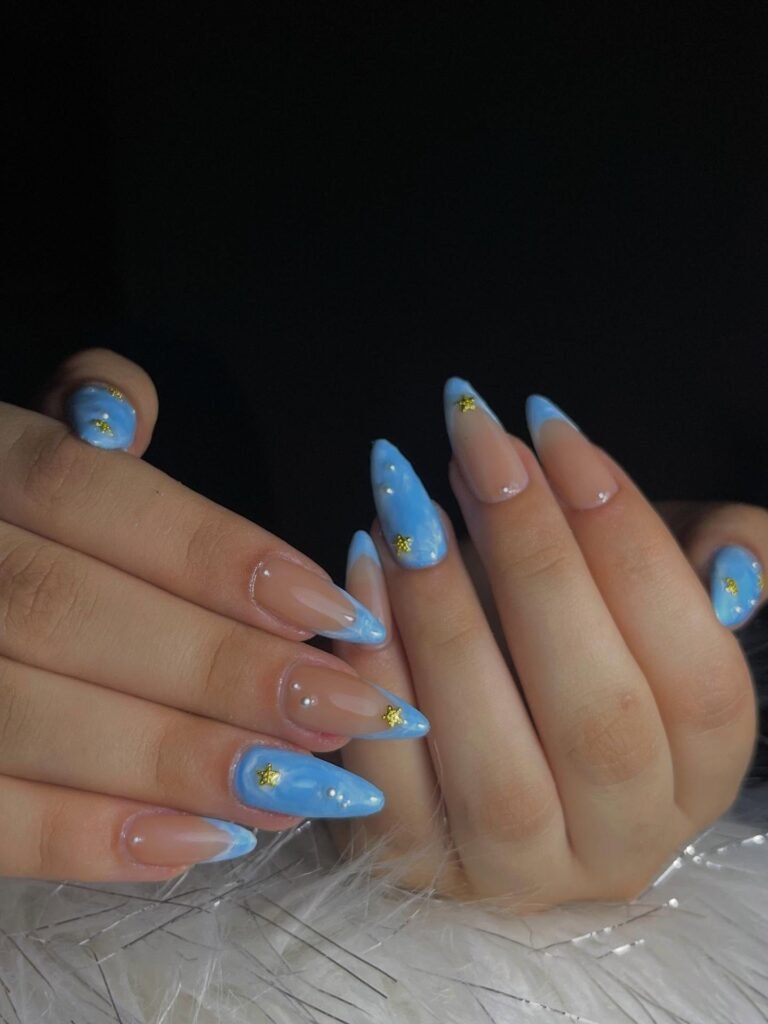 Blue and nude stiletto nails with pearl accents and yellow star details on hands, set against a dark background.