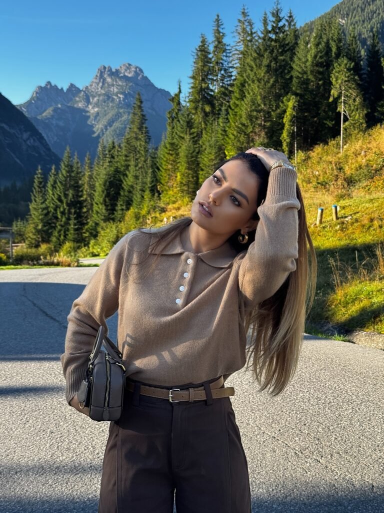 Woman in a brown sweater poses on a mountain road with fir trees in the background under a clear blue sky.