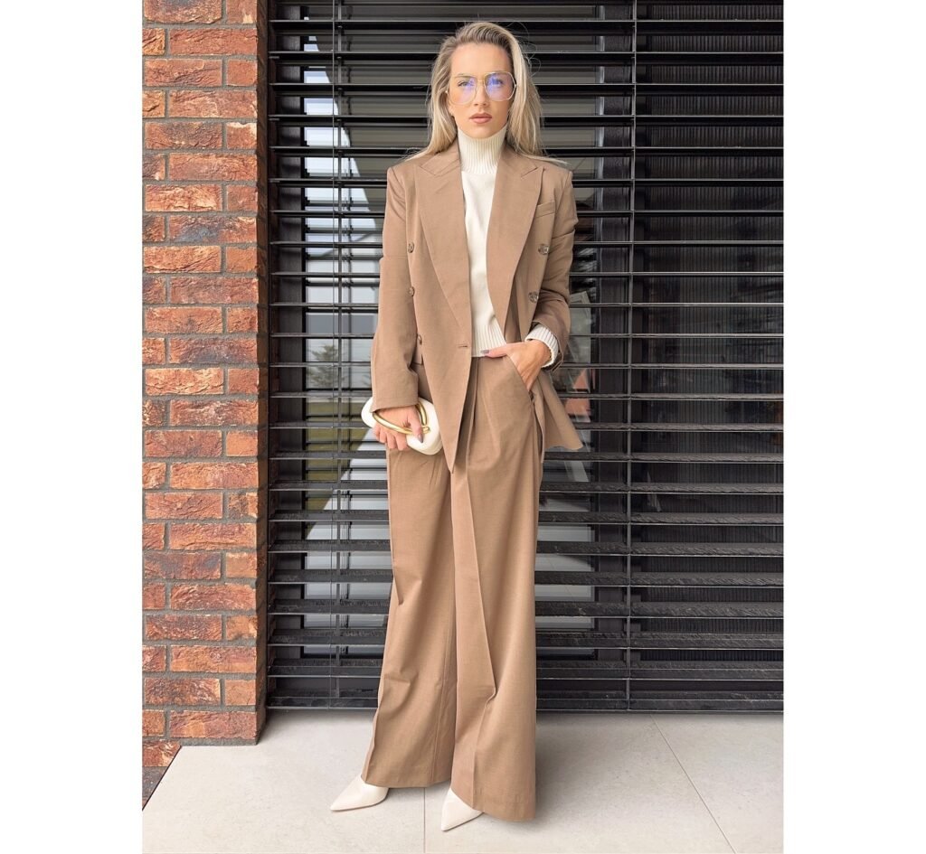 Woman in stylish brown tailored suit, white turtleneck; holding clutch, standing against brick and metal backdrop.
