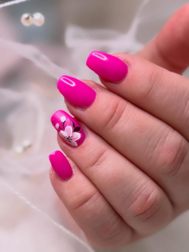 Bright pink nails with floral accent design on ring finger, showcasing stylish manicure on neatly shaped nails.