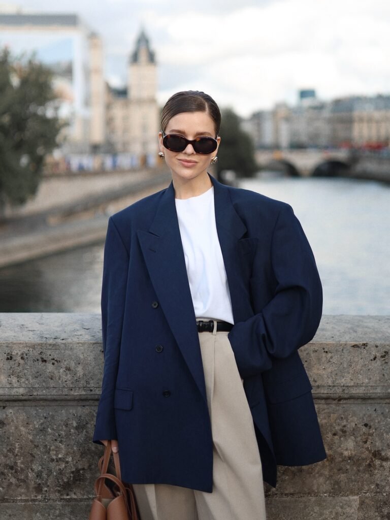 Fashionable woman in navy blazer and sunglasses standing by a river with elegant cityscape in the background.