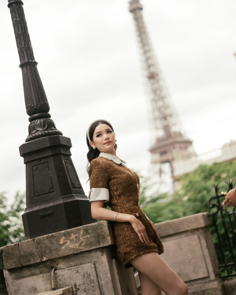Stylish woman poses near Eiffel Tower, Paris, in chic brown outfit, leaning against a decorative lamp post.