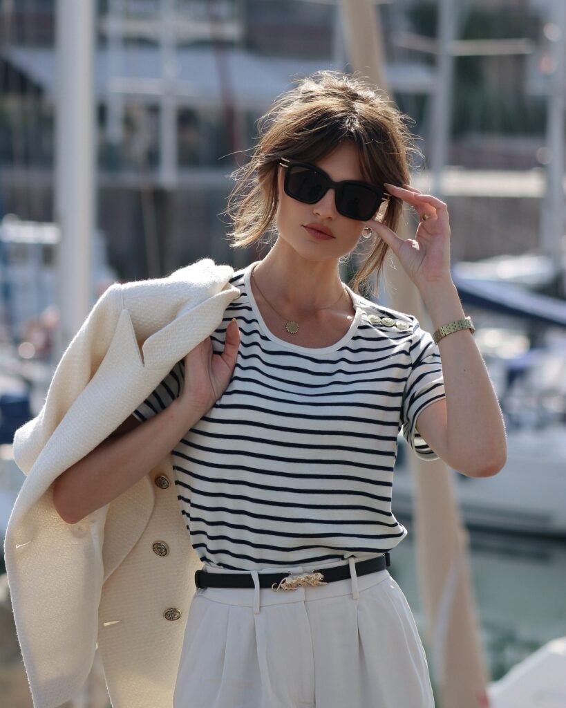 Stylish woman in striped shirt and sunglasses near marina, holding jacket over shoulder.