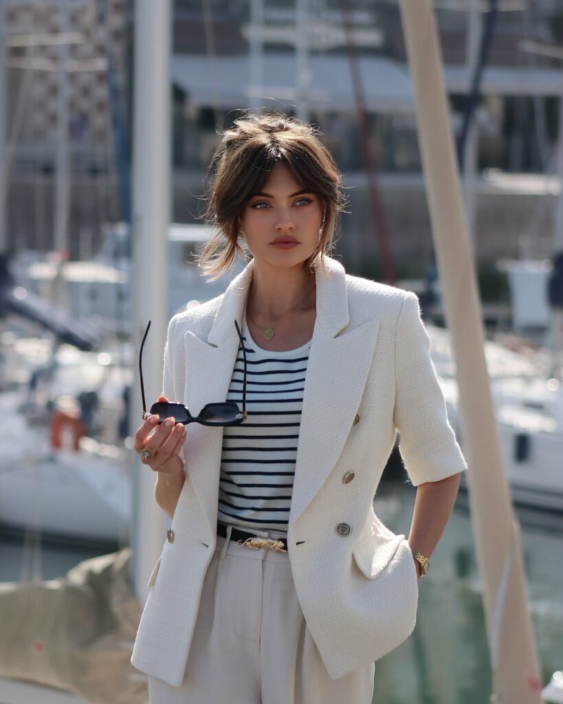 Stylish woman in nautical blazer and stripes by marina, holding sunglasses.