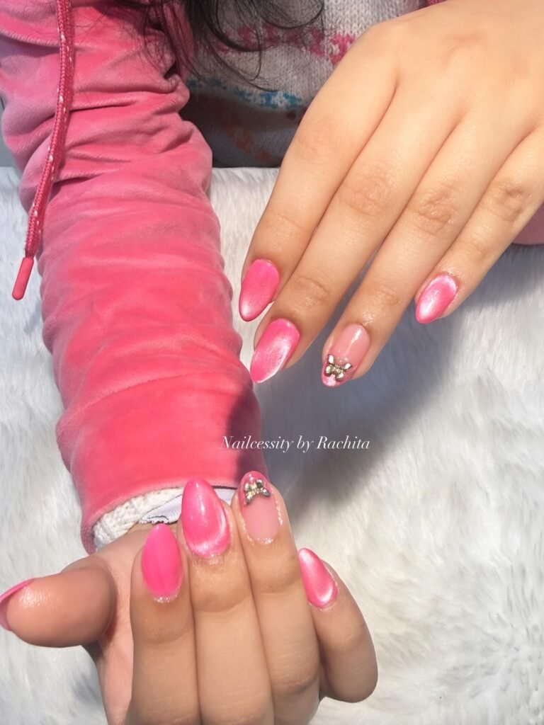 Pink nail art with rhinestone accents on a person’s hands, wearing a pink hoodie over a white fluffy surface.