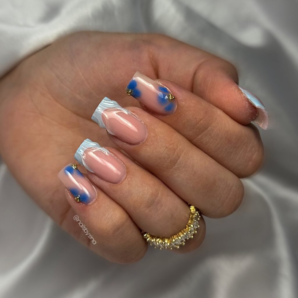 Close-up of hand with elegant pink and blue abstract nail art, adorned with gold accents, highlighting a jeweled ring.