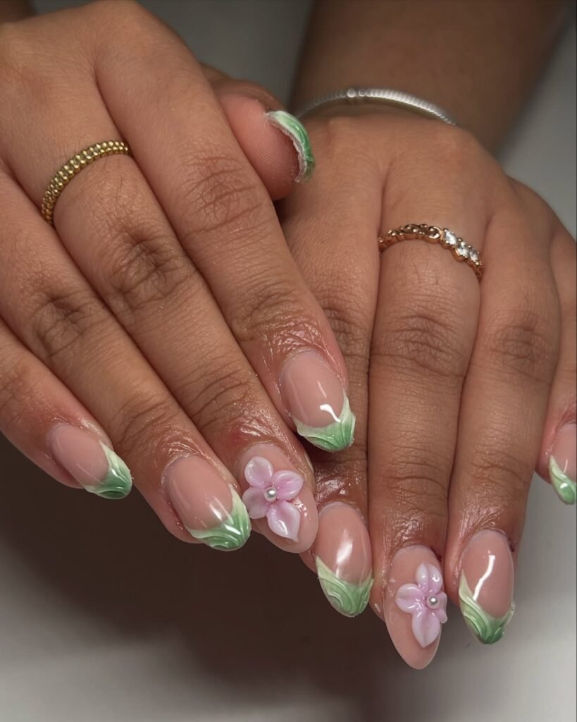 Nails with green leaf tips and 3D pink flowers, worn with gold and silver rings on both hands.
