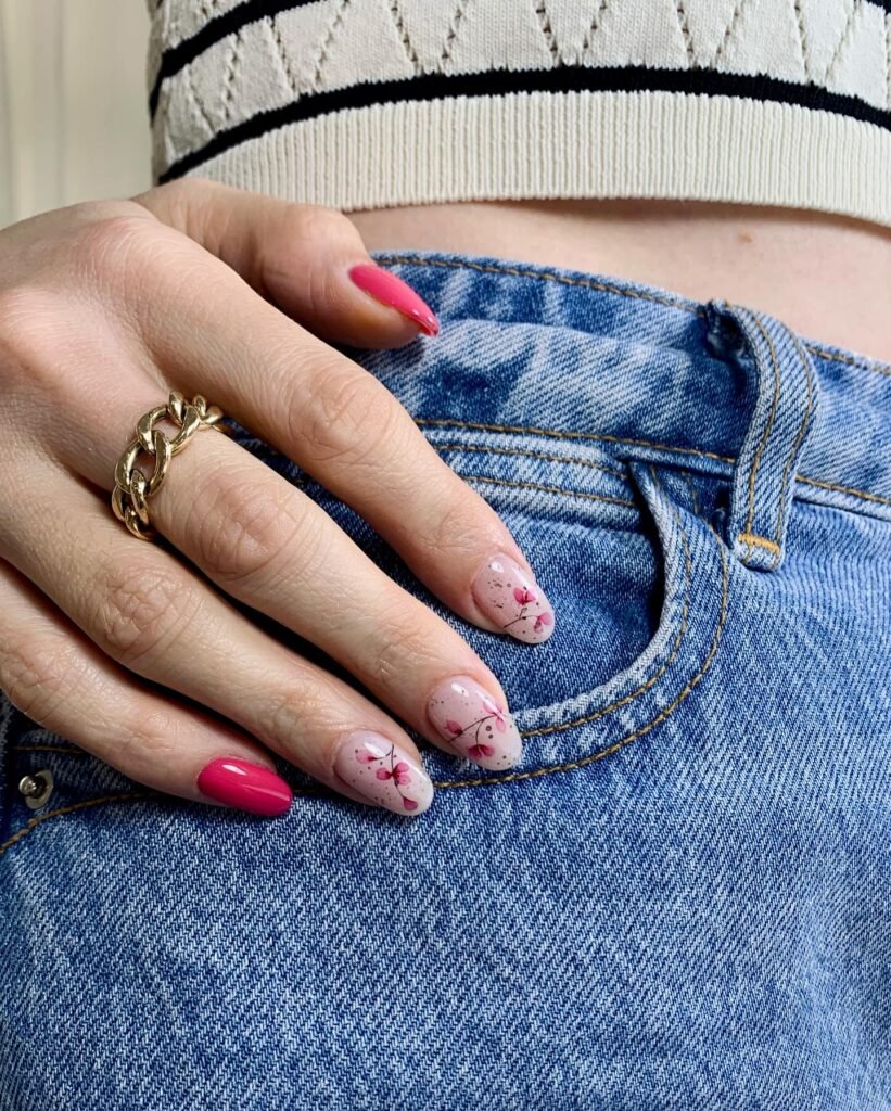 Hand with floral nail art and pink accent nail rests on blue jeans pocket, wearing a gold chain ring.