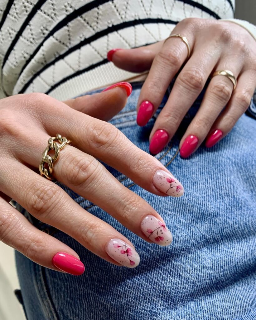 Close-up of hands with bright pink and floral nail art, wearing gold rings, resting on a denim surface.