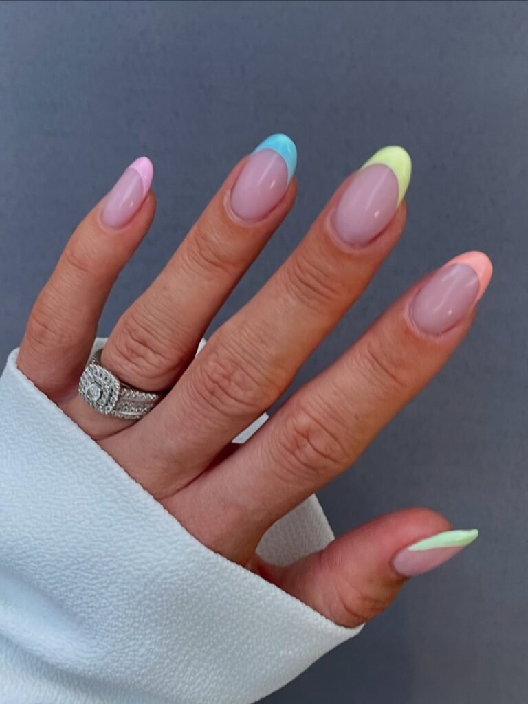 Hand with pastel French manicure and diamond rings, wearing a white sleeve, against a gray background.