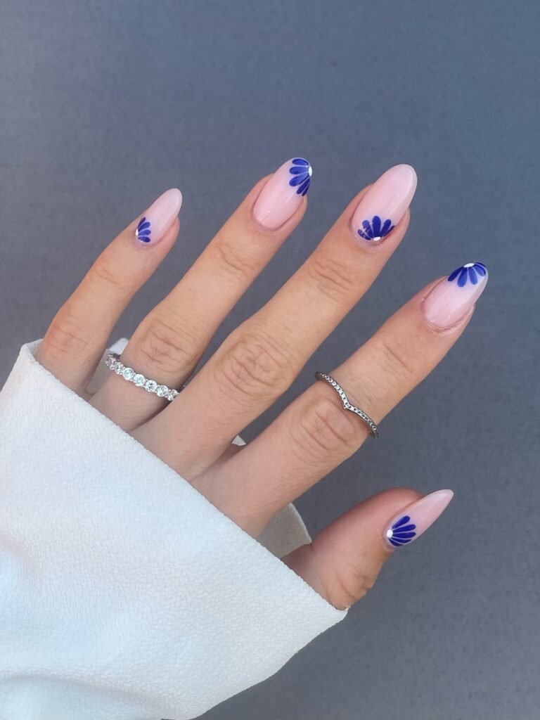 Elegant hand with pink nails featuring blue floral nail art, adorned with silver rings against a gray background.