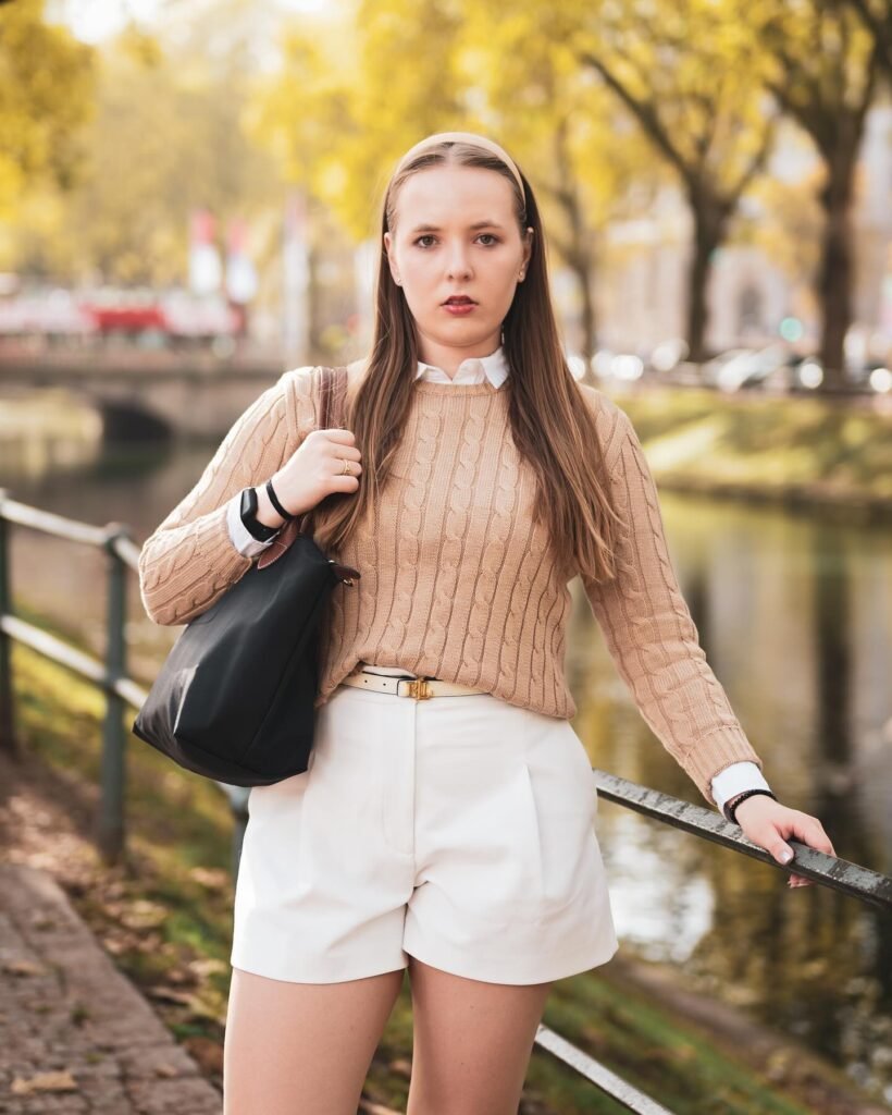 Young woman in a tan sweater and white shorts, standing by a park railing with autumn trees in the background.