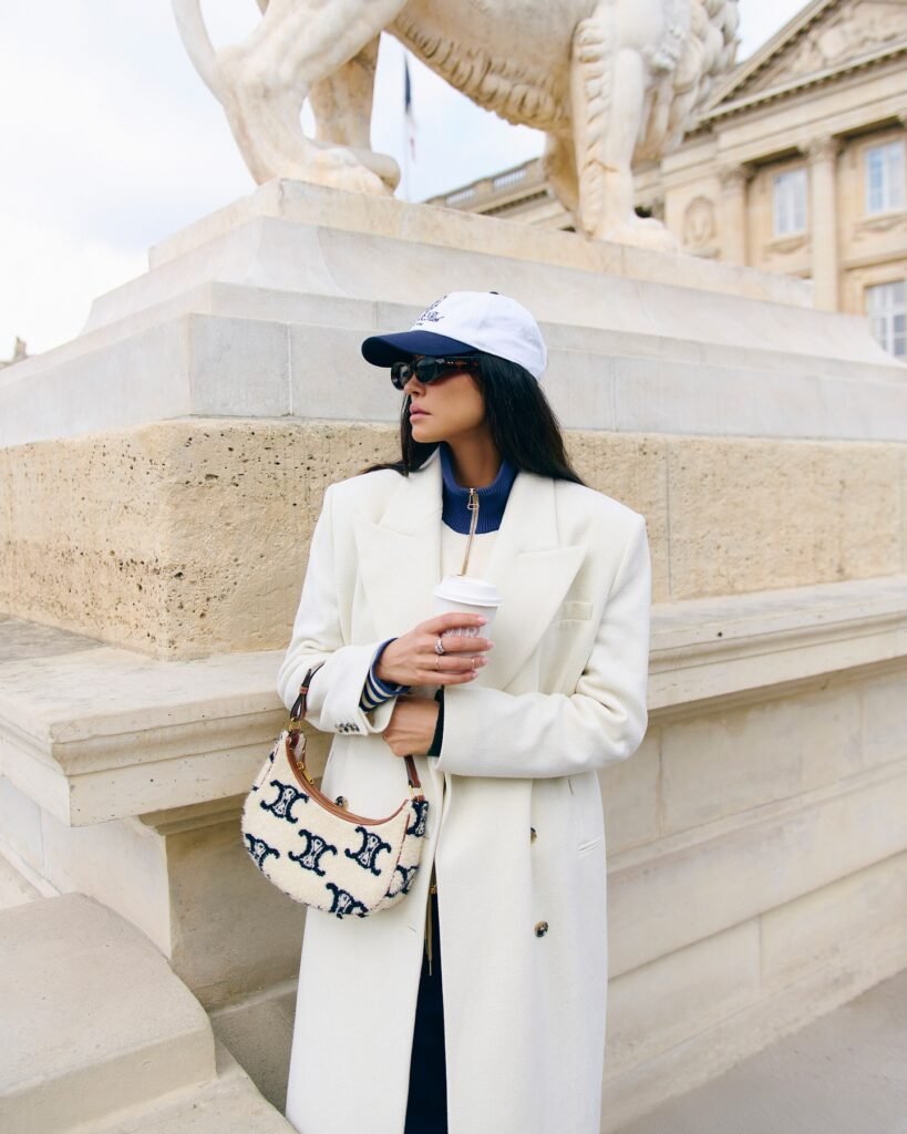 Stylish woman in white coat and cap holding coffee by a historical building and statue.