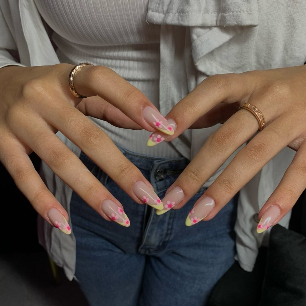 Stylish floral nail art design with pink flowers and yellow tips on manicured hands wearing gold rings.