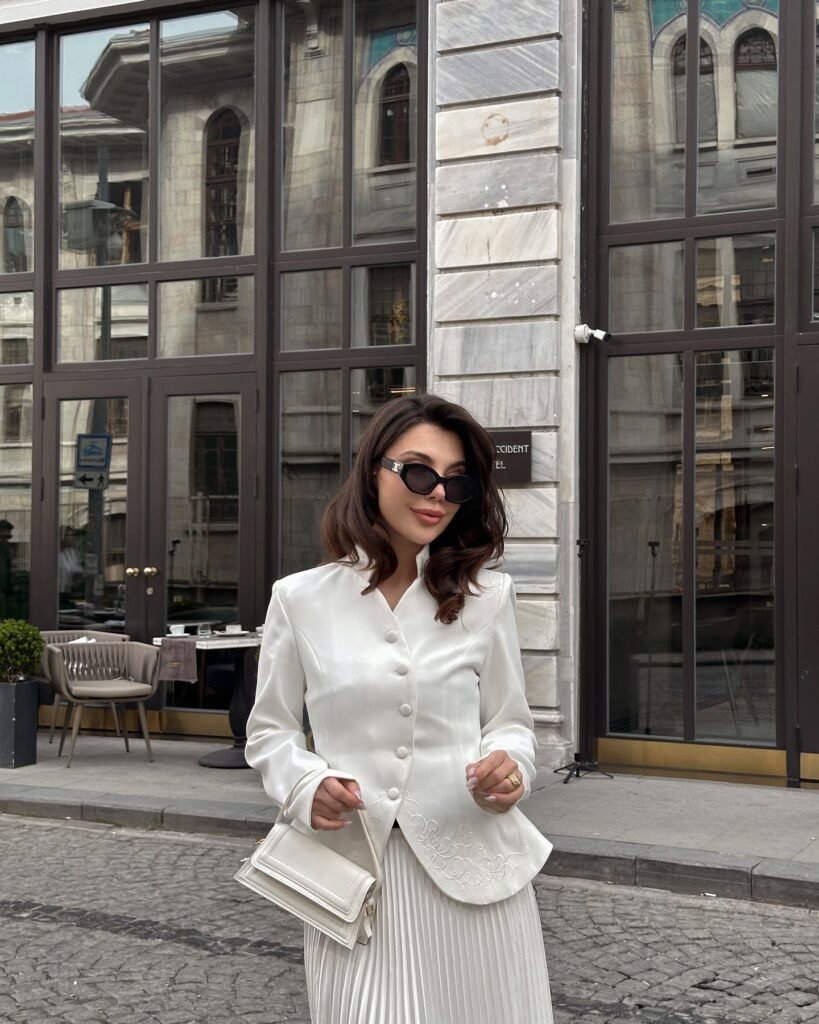 Woman in white outfit with sunglasses and handbag in front of modern building with large windows.