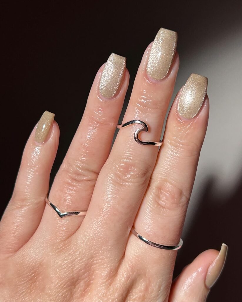 Elegant beige glitter manicure with silver rings on fingers.