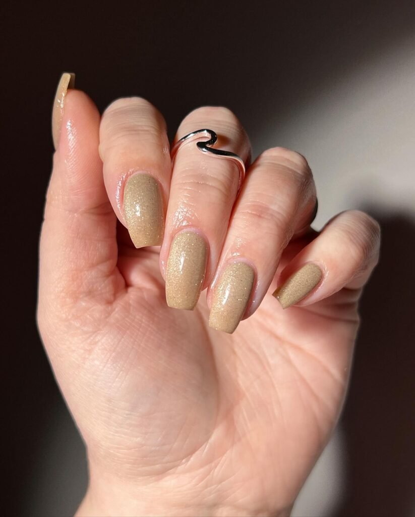 Close-up of a hand with glittery beige nail polish and a silver ring against a dark background.