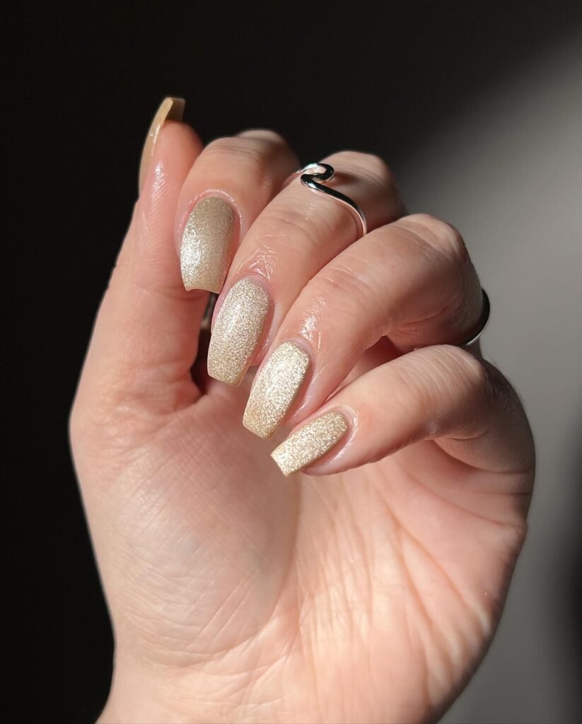 Glittery gold manicured nails with a silver ring on hand in soft lighting.