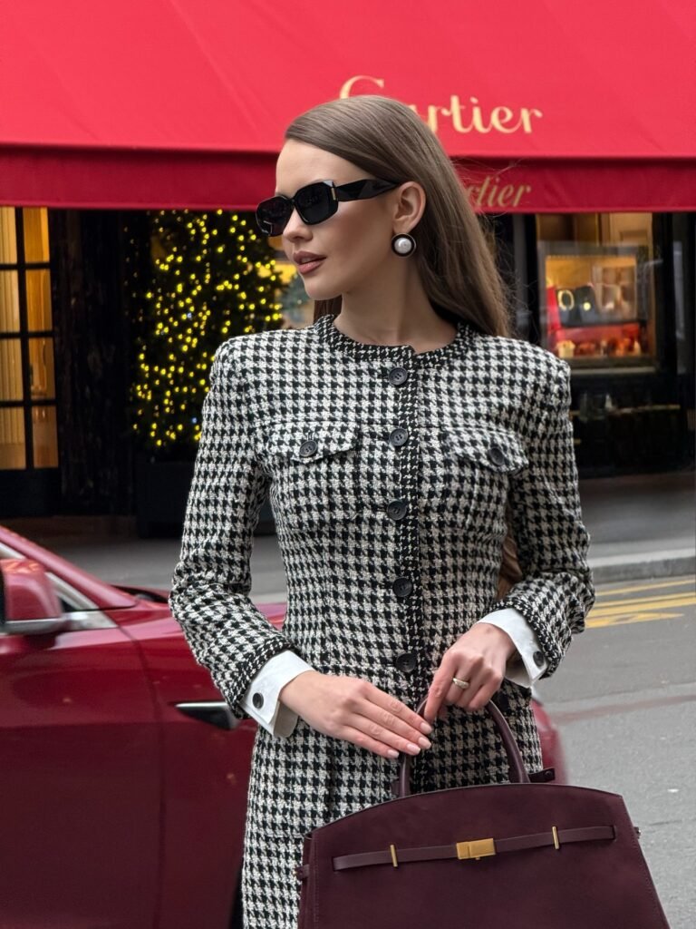 Stylish woman in houndstooth dress and sunglasses holding a burgundy handbag, standing in front of a luxury store.