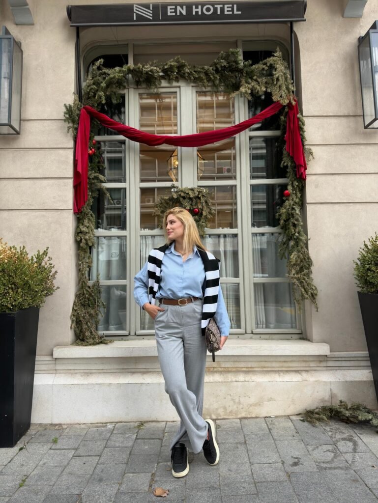Woman in stylish outfit poses by festive hotel window with wreath and garland decorations.